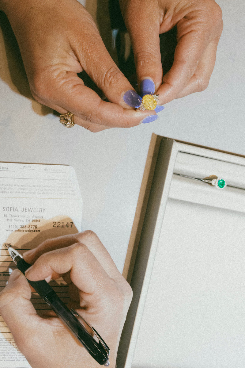 A woman holds a jewelry piece, thoughtfully designing her own wedding ring on a sheet of paper.